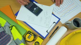 Building site surveyor working at his desk checking a spread sheet with a calculator, alongside a high visibility vest and a yellow hard hat