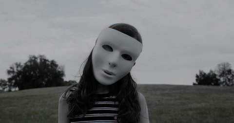 Young girl in white mask stands silently and creepily in a park