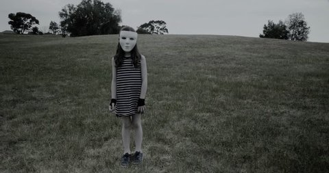 Young girl in white halloween masks tilts head looking creepy while standing in a park
