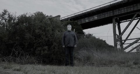 Creepy man wearing white halloween mask stands with head tilted in scary manner in front of bushes and old railway bridge
