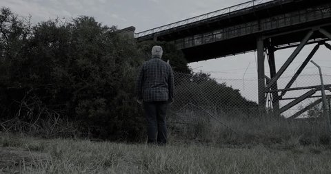 Creepy man wearing white halloween mask turns then stands with head tilted in scary manner then walks slowly away in front of bushes and old railway bridge