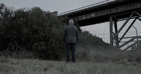 Creepy man wearing white halloween mask turns then stands with head tilted in scary manner in front of bushes and old railway bridge