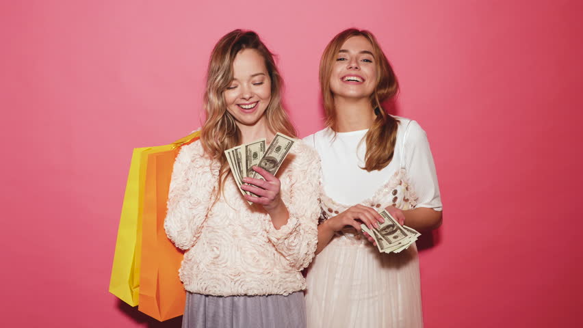Two young stylish smiling blond women holding shopping bags and making rain money. Hot girls dressed in summer hipster clothes and throwing bills out of a bundle money. Posing on pink background Royalty-Free Stock Footage #1017887587