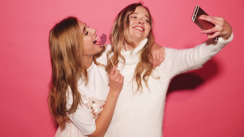 Two beautiful carefree girls taking selfie with a smart phone. Women make funny faces. Positive models posing over pink background with paper lips on stick. Female ready for party. 4k Royalty-Free Stock Footage #1017888118