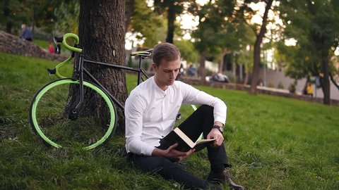 Young serious caucasian man reading book, sitting on the grass city urban park, studying or reading, leisure time in city park. Front view