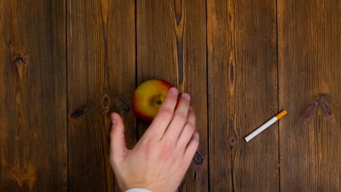 No smoking. Instead of smoking cigarettes eat fruits for healthy lifestyle. Man with his hand brushes cigarettes off away the wooden table and puts fresh apple. Make a choice or decision. Top view