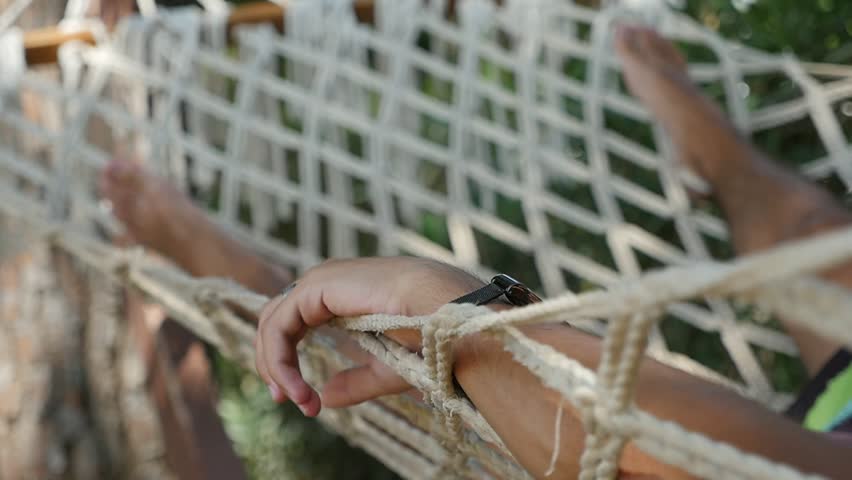 Funny view of young man`s legs lying carefree in a wide hammock woven from strong white ropes among trees on a sunny day in summer in slow motion