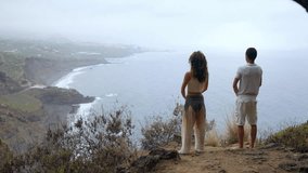 A man and a woman standing on the edge of a cliff overlooking the ocean raise their hands up and inhale the sea air during yoga