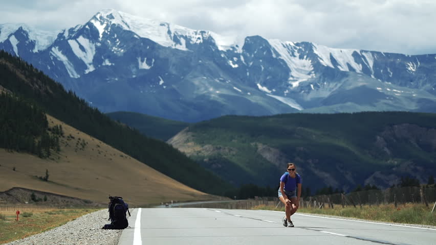 Young tourist woman with a backpack in shorts is sitting in the middle of the mountain road and is hitchhiking using two hands. There are snow mountains in the background. Long shot | Shutterstock HD Video #1017908968