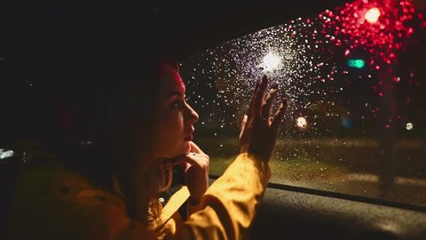Young Woman Looking Out of the Moving Car Window at Night. SLOW MOTION. Girl sitting on the back passenger seat of driving car. Night City Traffic on background.