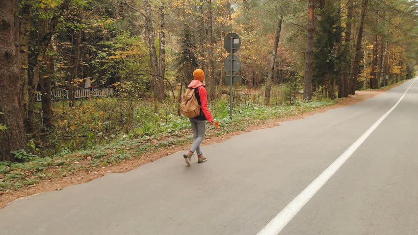 A young slim girl in a yellow hat and a vintage backpack walks along an asphalt road in the autumn yellow coniferous forest. Side back view