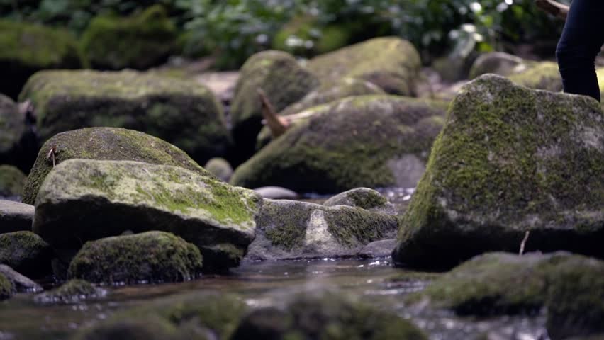 A young guy who is dressed as a hipster is jumping over a stream in the black forest. the camera is pointed at his shoes