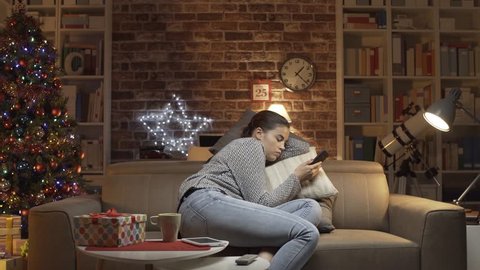 Sad woman having a lonely Christmas Eve at home on the sofa, she is checking her mobile phone but she is not receiving any message