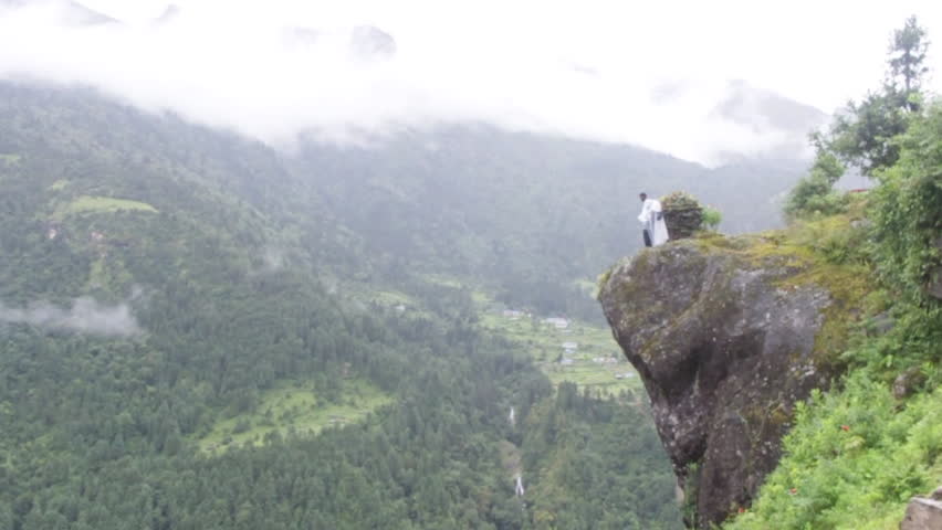 A man stands on a boulder overlooking a deep green valley. Green trees and low clouds make a dramatic view.