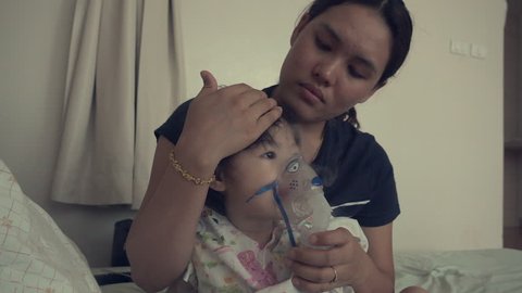 Little asian girl inhaling through inhaler mask with her mom at the hospital.