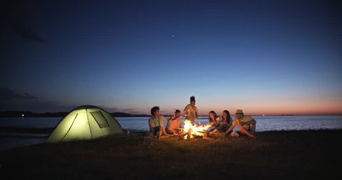 Group of six old multi-ethnic friends sitting near bonfire, roasting sausages, having a summer beach party, talking, enjoying their time 4k Stock Video