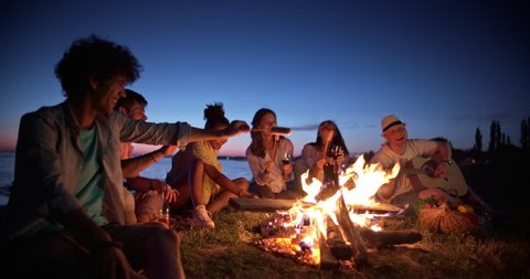 Group of six old multi-ethnic friends sitting near bonfire, roasting sausages, having a summer beach party, talking, enjoying their time 4k