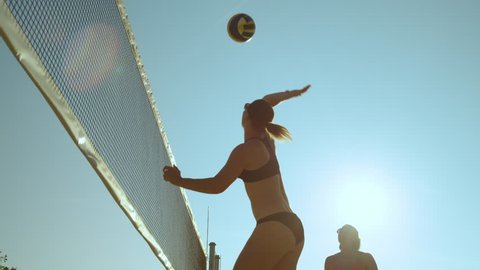 SLOW MOTION, CLOSE UP, LENS FLARE, LOW ANGLE: Athletic young Caucasian women in bikinis playing beach volleyball in the bright summer sun. Fit girls having fun playing volleyball in their bikinis.