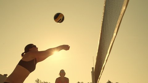 SLOW MOTION, LOW ANGLE, CLOSE UP, LENS FLARE: Young female volleyball players pass and spike the ball over the net on a sunny summer evening. Fit Caucasian girls playing beach volleyball at sunset.