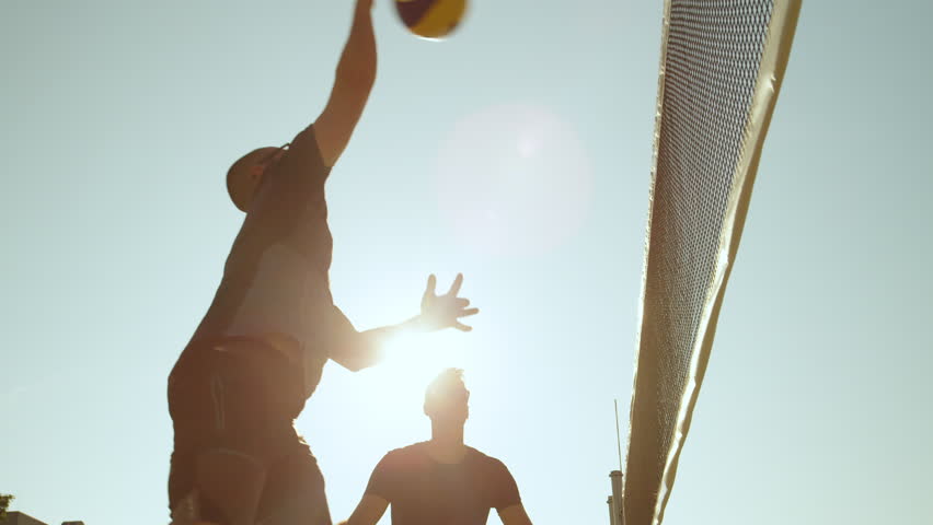 SLOW MOTION, CLOSE UP, LOW ANGLE, LENS FLARE: Athletic male voleyball player spikes the ball into the black net and fails to score a point. Fit Caucasian men playing beach volleyball during vacation.