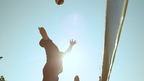 SLOW MOTION, CLOSE UP, LOW ANGLE, SUN FLARE: Athletic male playing beach volleyball spikes the ball over the net and scores a point. Fit guys playing volleyball during their fun summer vacation.
