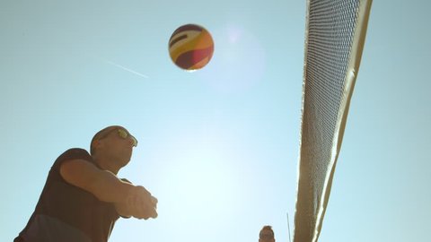 SLOW MOTION, CLOSE UP, LOW ANGLE, LENS FLARE: Adult male beach volleyball player strikes the ball over the net and scores a point. Two young guys on summer holiday playing volleyball on a sunny day. Stock Video