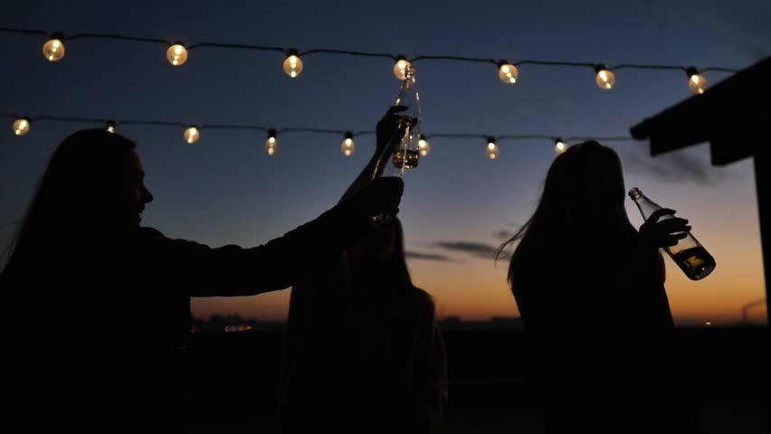Silhouettes of young people toasting with bottles and dancing with raised arms to the music played by dj at rooftop party during beautiful city sunset | Shutterstock HD Video #1017958993