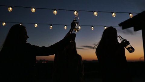 Silhouettes of young people toasting with bottles and dancing with raised arms to the music played by dj at rooftop party during beautiful city sunset