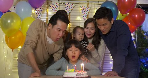 A cute little girl sitting at table and blowing candles on birthday cake while her family standing behind and sing a song to her. She gives a big smile and her family applaud her. 