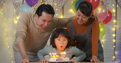 A cute little asian girl sitting at table and blowing candles on birthday cake while her family standing behind and sing a song to her. She gives a big smile and her family applaud her. 