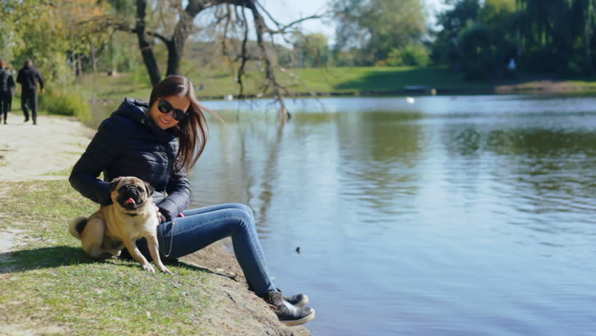 Young beautiful girl stroking her dog in a park near the lake. Dog and owner sitting on the ground