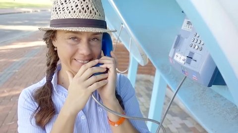 adult funny woman is talking by phone in call box on a street in summer day, looking at camera