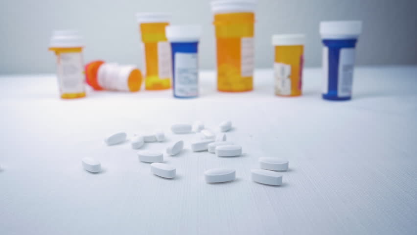 Prescription pill bottle falls on a pile of narcotics in slow motion. Pharmaceutical bottles sit in the background. The painkillers represent the opioid crisis in America, addiction and overdoses. Royalty-Free Stock Footage #1017973762