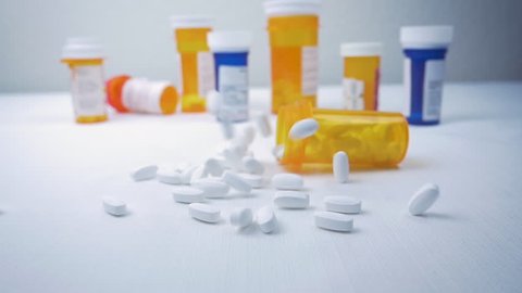 Prescription pill bottle falls on a pile of narcotics in slow motion. Pharmaceutical bottles sit in the background. The painkillers represent the opioid crisis in America, addiction and overdoses.