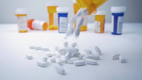 Prescription pills fall into a pile from a bottle in slow motion. Pharmaceutical bottles sit in the background. Painkillers represent the opioid crisis in America, narcotic addiction and overdoses.