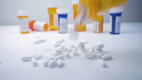 Bottle of painkillers falls on a pile of white narcotic pills in slow motion.  Prescription bottles clutter the background. Capsules represent the opioid crisis in America and narcotic addiction.
