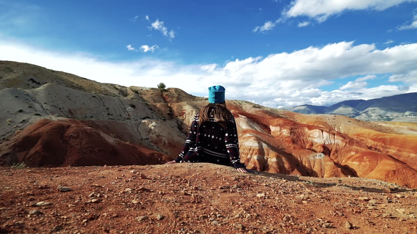 Young woman tourist sits on the edge of a red mountain. There are scenic landscape and cloudy sky in the background. Back view, slider