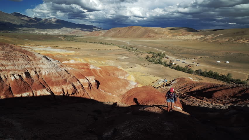 Young tourist woman climbs a red stone mountain in sunny day. There are scenic landscape and cloudy sky in the background