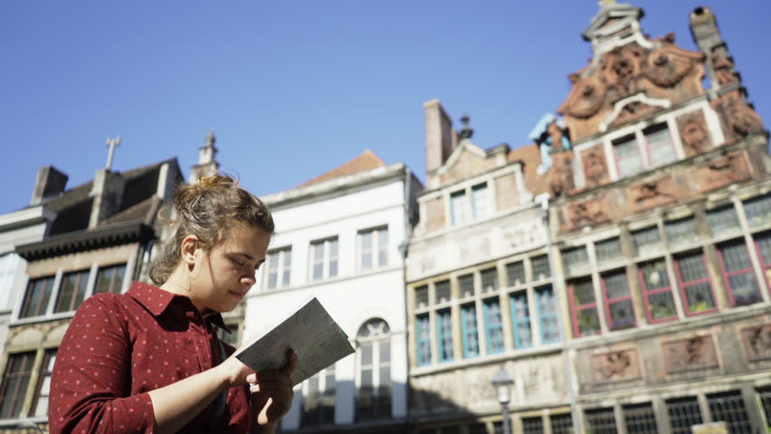 Girl is walking along the touristic center of Ghent on a sunny day. Bottom shot Worried woman with map in hands at blurred background of traditional Belgian buildings and blue sky