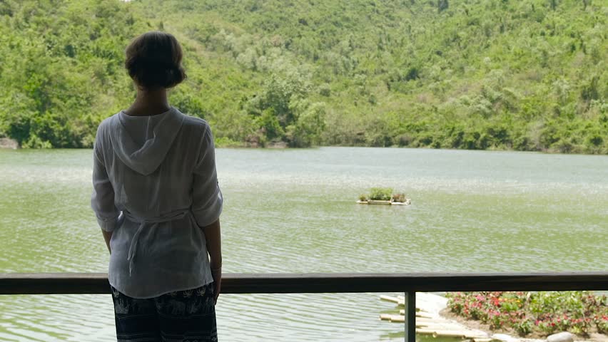 Young woman standing on background river and mountain covered green forest. Girl watching on river and green mountain landscape, back view