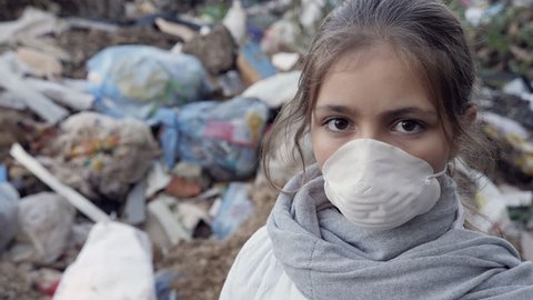 Young girl in a white respirator at the garbage dump looks into the camera. Ecology concept. Slow motion video