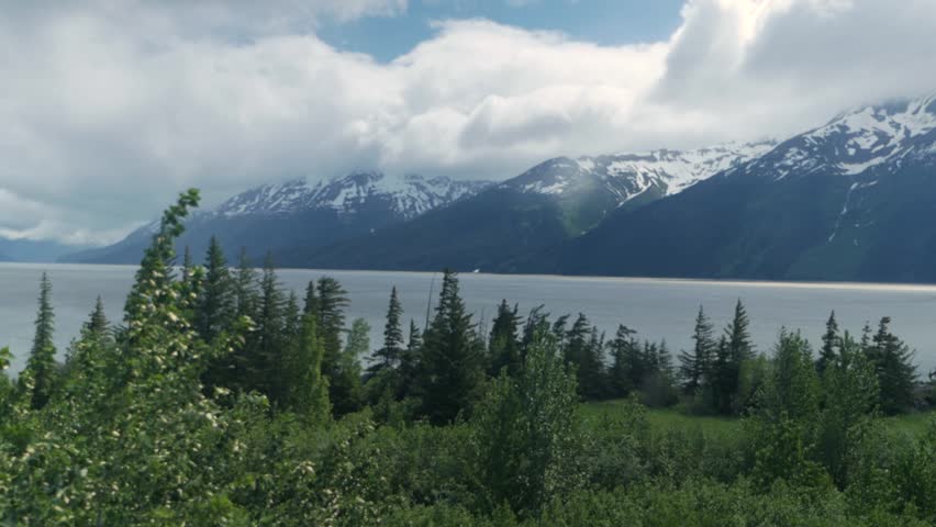 Alaska Railroad passenger train travels along coastline with snow-capped mountains on the horizon.