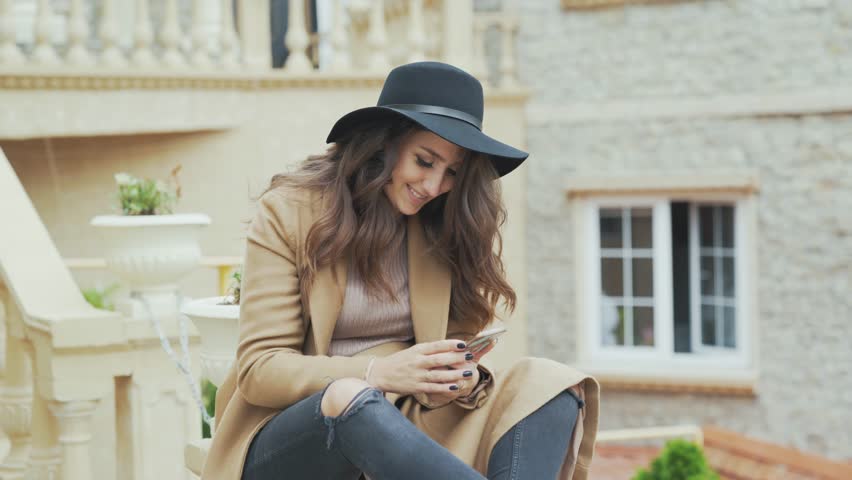 Attractive smiling European with curly hair, wearing a stylish hat and coat, talking to someone on her smartphone, making an appointment, writing SMS from her phone.