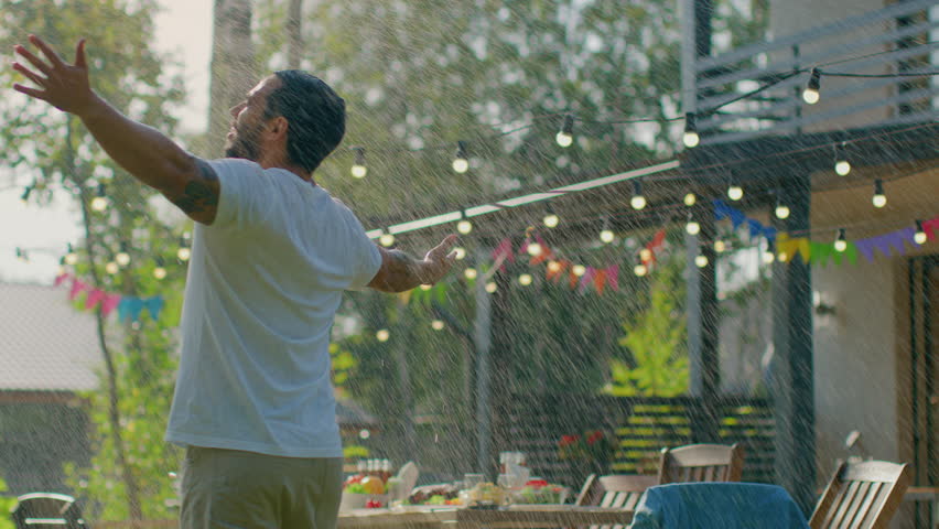 Handsome Fit Young Man Spins and Dances Under the Rain Made by Garden Water Hose Sprinkler, Raises Arms. Muscular Guy Cools on a Hot Summer Day. Royalty-Free Stock Footage #1018014964