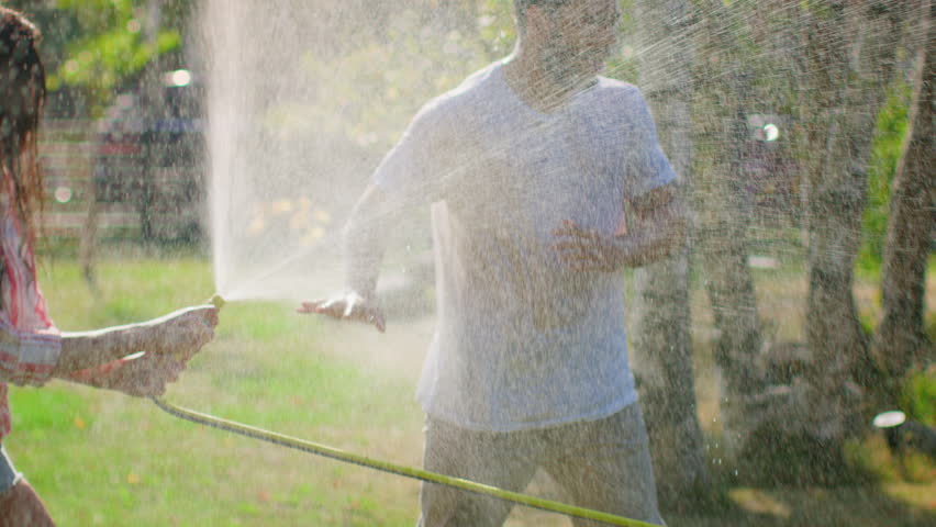 Happy Young Couple Has Fun on a Hot Summer Day Playing with Water Hose Sprinkler in the Garden. Two Young People in Love Got Wet Jokingly Fighting with Hose. In Slow Motion. Royalty-Free Stock Footage #1018015867