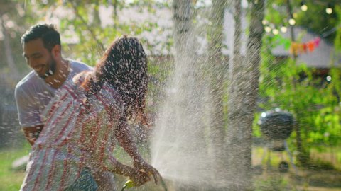 Happy Young Couple Has Fun on a Hot Summer Day Playing with Water Hose Sprinkler in the Garden. Two Young People in Love Got Wet Jokingly Fighting with Hose. In Slow Motion.