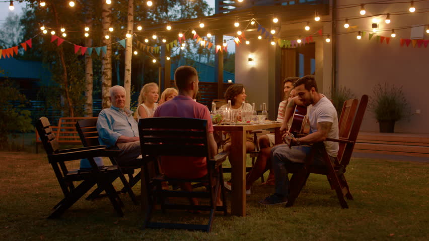 Sitting at the Dinner Table Handsome Young Man Plays the Guitar For a Friends. Family and Friends Listening to Music at the Summer Evening Garden Party Celebration. Royalty-Free Stock Footage #1018015894