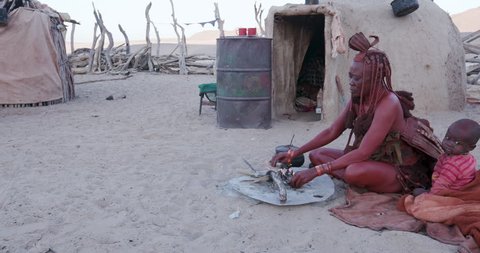 4K view of Himba woman in traditional dress with young child, putting a small pot on a fire outside their hut within their small compound, Namibia