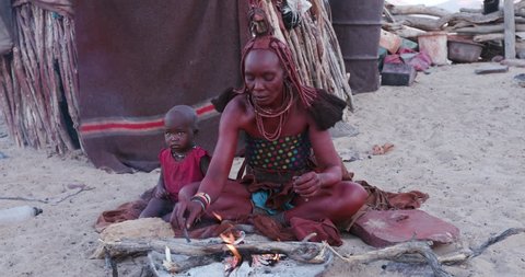 4K side view of Himba woman in traditional dress with young child, filling a pipe and smoking outside their hut within their small compound, Namibia