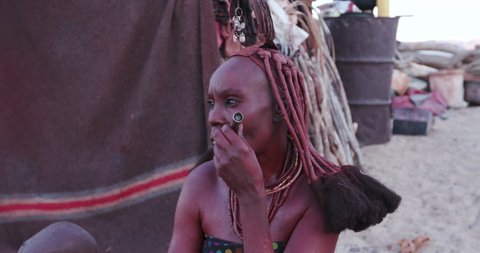 4K close-up view of Himba woman in traditional dress with young child, smoking a pipe outside their hut within their small compound, Namibia